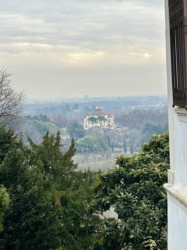 A scenic view featuring a distant villa surrounded by trees and hills under a cloudy sky.