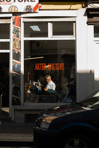 Picture of a tailoring shop. A worker inside is looking towards the camera while working with a sewing machine.

Foto di una sartoria, dove il sarto che sta lavorando con una macchina da cucire accanto alla finestra guarda fuori verso la camera.