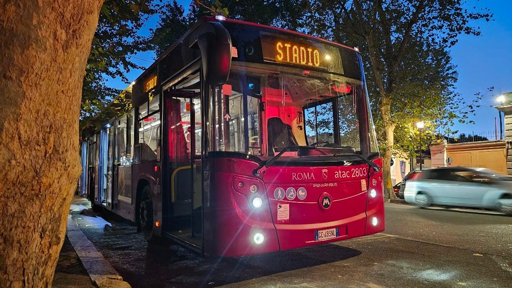 La fotografia mostra un autobus urbano Atac di colore rosso scuro e grigio, fermo lungo una strada alberata nelle ore serali. Sul display luminoso anteriore compare la destinazione “STADIO”, mentre sulla parte frontale sono visibili il logo Atac e il numero del veicolo. I fari sono accesi e illuminano la carreggiata. Un grande tronco d’albero occupa il lato sinistro dell’immagine; sullo sfondo si vedono alberi, edifici e un’automobile in movimento. La scena documenta un autobus predisposto per il collegamento con lo stadio in occasione dell’evento sportivo.