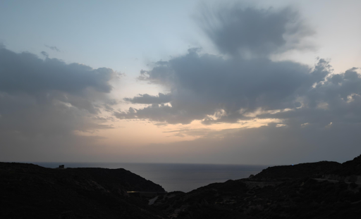 Il tramonto mostra un cielo sfumato dal celeste chiaro fino a tonalità calde e aranciate vicino all'orizzonte. Grandi nuvole grigie e dense coprono gran parte del cielo, lasciando filtrare la luce soffusa del sole calante. In basso, il mare calmo è incorniciato dal profilo scuro e collinare della costa in controluce.