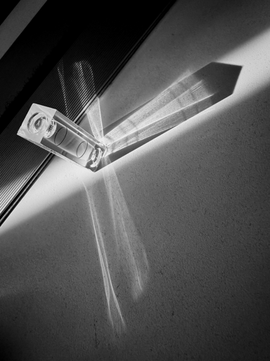 La foto in bianco e nero mostra un piccolo livello a bolla trasparente appoggiato su una superficie liscia. La luce del sole entra da sinistra, creando un’ombra nitida e riflessi luminosi sul piano. Il vetro del livello proietta linee e fasci di luce incrociati, dando un effetto geometrico e astratto.