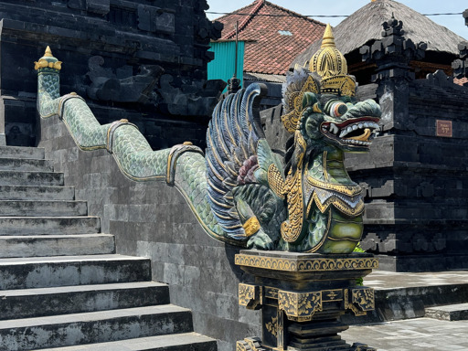 Ornately decorated nāga guardian beside a stone staircase near Tanah Lot Temple in Bali, Indonesia, photographed in August 2024. The mythical serpent-like creature has a long scaled green body winding up the staircase, a fierce open-mouthed face, feather-like wings and an elaborate golden headdress. Its vivid green, blue, white and gold details stand out against the dark volcanic-stone architecture of the surrounding Balinese temple complex.