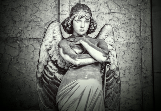 Un’immagine fotografica in bianco e nero raffigura una statua posizionata contro un muro di pietra texturizzato. La statua presenta ali grandi, ricoperte di piume, un cinturino, capelli ondulati e un drappo che cade. Le braccia sono incrociate sul petto. Il muro di pietra dietro ha un aspetto mottled e fratturato con divisioni verticali visibili. Il tono monocromatico enfatizza le texture della pietra e i dettagli della statua.