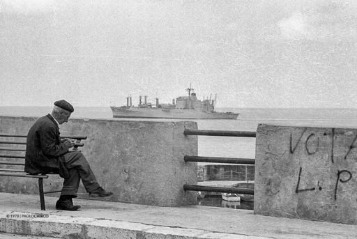 Un vecchio seduto su una panchina a Terrasini mentre rolla una sigaretta, indifferente alla nave militare ed ai suoi commerci
