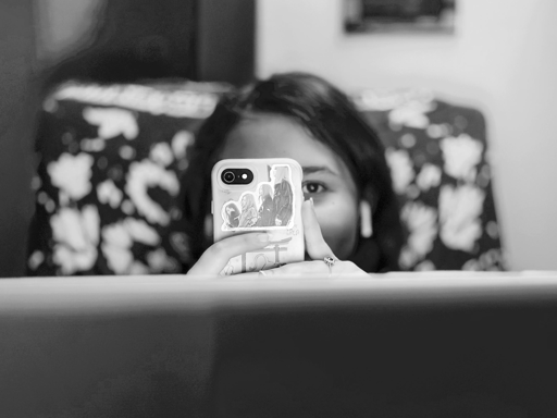 Questa è una fotografia in bianco e nero che ritrae una persona parzialmente nascosta dietro uno smartphone e lo schermo di un computer portatile, creando un'atmosfera di mistero e quotidianità digitale.

Composizione e Soggetto

• In primo piano: La parte inferiore dell'immagine è occupata dal retro sfocato dello schermo di un laptop, che funge da barriera visiva.
• Al centro: Una persona tiene in mano uno smartphone, posizionato proprio davanti al viso. Del volto sono visibili solo gli occhi e la fronte; uno degli occhi guarda direttamente verso l'obiettivo con un'espressione neutra ma attenta.