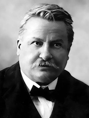 Ritratto fotografico in bianco e nero del poeta Giovanni Pascoli. Capelli corti, brizzolati, con la riga a destra, e baffi che coprono il labbro; viso rotondo, occhi che guardano in alto, e sguardo serio. Indossa una giacca scura con camicia bianca e cravattino a farfalla nero