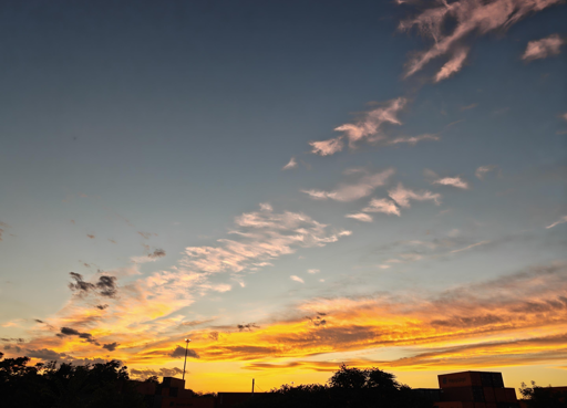 Questo tramonto si distingue per un forte contrasto cromatico e un'interessante disposizione delle nuvole.Colori e LuceOrizzonte infuocato: tonalità giallo vivo e arancione intenso.Cielo superiore: sfumature sfumate dal blu avio al ceruleo.Nuvole illuminate: riflessi rosa confetto e dorati sui bordi.Nuvole e AtmosferaFormazione diagonale: una scia di nuvolette sfilacciate attraversa il cielo.Tonalità miste: nuvole scure in basso e nuvole cariche di luce in alto.Effetto dinamico: il vento sembra muovere i cirri verso l'alto.Elementi in OmbraProfilo urbano: silhouette nere di edifici e alberi sul fondo.Palo della luce: un sottile elemento verticale taglia la linea dell'orizzonte.