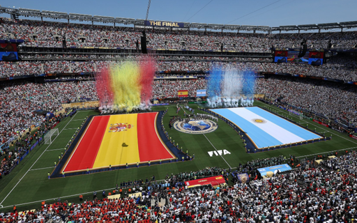Una vista panoramica dall'alto di un grande stadio di calcio pieno di spettatori durante una finale. Sul campo da gioco sono distese due grandi bandiere: una con strisce rosse e gialle e l'altra con strisce azzurre e bianche. Colonne di fumo colorato in rosso, giallo, azzurro e bianco si elevano sopra le aree coperte dalle bandiere. Le tribune sono gremite di persone e la scritta "THE FINAL" è visibile nella parte superiore centrale della struttura dello stadio. Un logo circolare e il testo "FIFA" sono visibili sul prato tra le due bandiere.