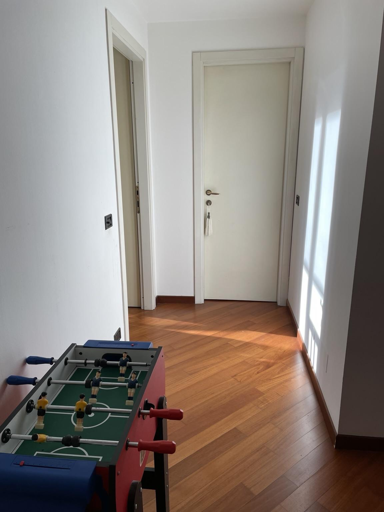 L'immagine mostra un corridoio con pavimento in legno di colore marrone chiaro, che riflette la luce naturale proveniente da una finestra a destra. Le pareti sono dipinte di bianco, creando un'atmosfera luminosa e spaziosa. In primo piano, a sinistra, si trova un tavolo da calcio, con manopole rosse e blu e figure di giocatori in giallo e blu, pronto per un gioco. In fondo al corridoio, si vede una porta bianca con una maniglia e una tassella, che sembra essere chiusa. A sinistra, c'è un'altra porta aperta che conduce a un'altra stanza. L'ambiente è semplice e minimalista, con un'attenzione particolare alla pulizia e all'ordine.