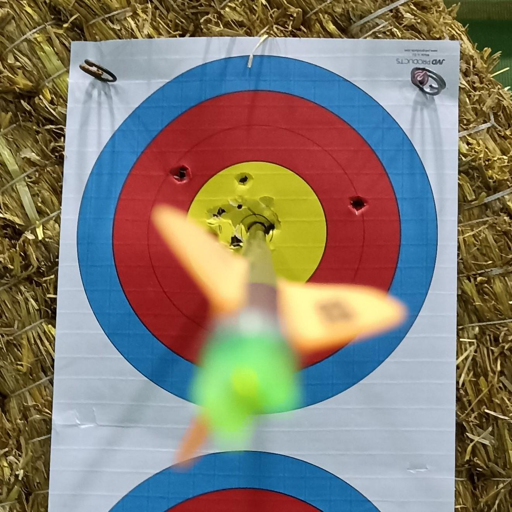 Un bersaglio da tiro con l'arco è appeso su uno sfondo di paglia. Il bersaglio è circolare e diviso in cinque cerchi concentrici: giallo, rosso, blu, blu scuro e bianco. Sono visibili diversi fori nel bersaglio, principalmente nel cerchio giallo. Una freccia ha fatto in centro perfetto.