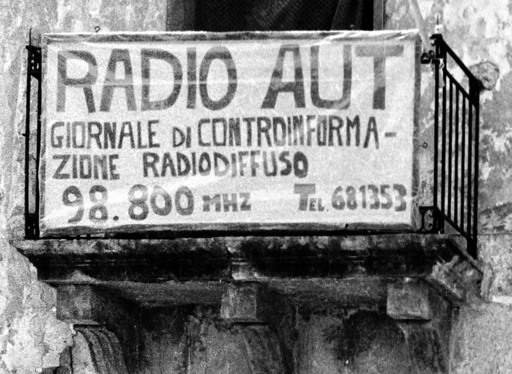 Fotografia bn del pannello sul balcone della sede della radio con scritta: radio aut giornale di controinformazione radiodiffuso 98.800Mhz tel. 681353