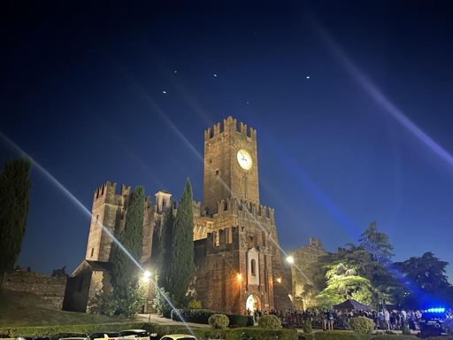 A medieval castle illuminated at night, featuring a tall tower with a clock. Surrounding greenery includes tall trees, and a crowd is gathered near the entrance, with additional lighting effects in the sky.