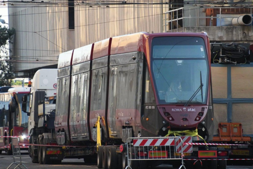 La fotografia mostra un tram CAF Urbos con livrea rosso scuro e grigia, caricato su un autoarticolato per il trasporto su strada. La parte anteriore del convoglio è rivolta verso chi osserva: si distinguono il grande parabrezza, i fari, il logo Atac e la scritta Roma. Il tram è fermo in un’area di servizio o di deposito, circondato da mezzi pesanti e attrezzature. Sullo sfondo si vedono edifici industriali e cavi della rete tranviaria aerea. Barriere mobili e segnalazioni delimitano lo spazio operativo intorno al veicolo.