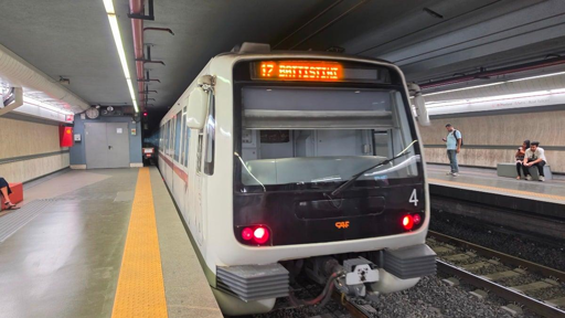 Un treno della metropolitana è fermo in una stazione sotterranea. Il convoglio, bianco con fascia rossa e dettagli scuri, è ripreso frontalmente dal marciapiede. Sul display anteriore è visibile la destinazione della corsa. Alcuni passeggeri attendono sul marciapiede, mentre la banchina presenta la consueta fascia tattile gialla lungo il bordo.