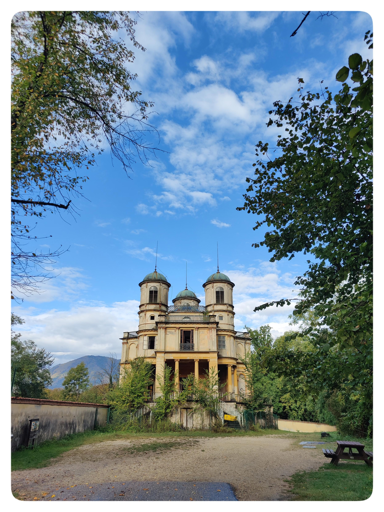 La Bizzarria.
Una strada sterrata (nella parte bassa della foto) porta al casino di caccia. La struttura ha due torrette laterali e una più piccola centrale. Il colore è un classico giallo Piemonte (o sabaudo o Torino).
Il cielo sereno incanta con il suo azzurro splendente e le bianche nuvole che portano lo sguardo alle montagne in lontananza.