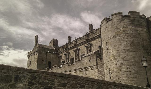 Fotografia che ci mostra il castello di Stirling sotto un cielo nuvoloso. L'inquadratura dal basso sottolinea la poderosa torre rotonda e le merlature che sovrastano il cortile, ma mostra anche le quattro nicchie con statue sulla facciata, che ne attestano la ricchezza delle decorazioni