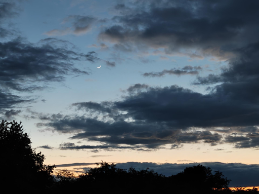 Il cielo al crepuscolo si tinge di sfumature delicate e contrasti decisi. Un orizzonte luminoso sfuma dal giallo tenue all'arancione pallido. Il cielo vira verso un blu profondo e freddo. Masse nuvolose grigie e dense coprono gran parte della scena. Lembi leggeri di fumo scuro fluttuano vicino ai corpi celesti. Una sottile falce luminosa brilla tra le nuvole nel cielo blu. Un piccolo punto luminoso punge l'oscurità poco sopra la Luna. La vegetazione in basso appare come una linea d'ombra completamente scura. Un vivo riflesso arancione si intravede tra i rami sul margine destro.