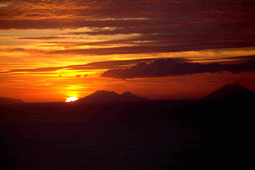 Foto di un tramonto. Colori caldi, il cielo è nuvoloso ed il sole si trova nel primo terzo a sinistra, quasi del tutto tramontato, coperto parzialmente da una montagna, presumibilmente lo Stromboli.