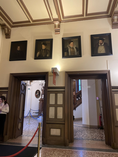 L'immagine mostra un corridoio interno decorato con elementi architettonici classici. Quattro ritratti incorniciati sono disposti in fila orizzontale sulla parete superiore. Sotto i ritratti ci sono due porte in legno scuro, ognuna con pannelli decorativi. La porta a sinistra conduce a uno spazio illuminato visibile, mentre quella a destra è chiusa e rivela un'altra stanza parzialmente illuminata. Al centro tra le due porte c'è una colonna decorativa. Sulla parete sinistra è visibile la scritta "ENTRATA".
