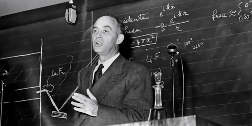 Fotografia in bianco e nero di Enrico Fermi. La foto sembra scattata durante una lezione o una conferenza; Fermi sta parlando e gesticola con la mano sinistra, mentre sta di fronte ad una lavagna con alcune formule scritte, rivolto in direzione di un microfono. Veste in giacca e cravatta.