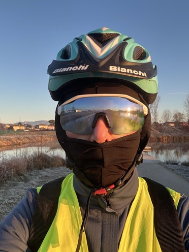 A person in a cycling helmet and sunglasses is wearing a black face mask and a yellow reflective vest. The background features a frosty path, water, and trees under a clear sky during sunrise.