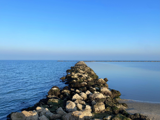 This image captures a serene, wide-angle coastal scene featuring a rock breakwater (or jetty) stretching out into a calm sea under a clear blue sky.
Composition and Subject
• The Foreground: A path of jagged, uneven light-colored rocks leads the viewer's eye from the bottom of the frame toward the horizon. Some rocks are covered in dark green moss or seaweed, suggesting the ebb and flow of the tide.
• The Water: The sea is divided by the jetty. To the left, the water shows gentle ripples and a deeper blue hue. To the right, the water is exceptionally still, acting like a mirror that reflects the soft gradient of the sky.