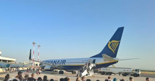 La fotografia mostra un aereo passeggeri della compagnia Ryanair fermo sulla pista in un’area aeroportuale. Il velivolo, con fusoliera bianca e blu e logo giallo sulla coda, è circondato da passeggeri in attesa e personale a terra. Una scala mobile permette l’accesso all’aereo. Sul lato destro si vede un’autocisterna per il rifornimento. Sullo sfondo sono visibili edifici aeroportuali e il cielo sereno. L’immagine accompagna la notizia sulle limitazioni al traffico aereo causate dal guasto ai sistemi di rete di ENAV.