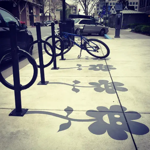 L'immagine mostra una scena urbana con un marciapiede che ospita un sistema di parcheggio per biciclette. Al centro dell'immagine, un mountain bike blu è appoggiato a un paliere di metallo nero, che presenta cerchi per fissare le biciclette. Il marciapiede è decorato con un'immagine dipinta a terra di fiori e foglie, che proiettano ombre chiare sul cemento. Sullo sfondo, si vedono auto parcheggiate lungo la strada e edifici urbani. L'atmosfera è tranquilla, con un cielo nuvoloso che diffonde una luce soffusa.