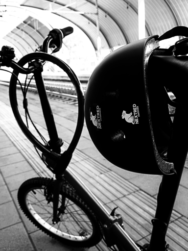 Una foto in bianco e nero, una bici pieghevole parcheggiata. Di sfondo una stazione nuova suburbana. In primo piano un caschetto da bici, con il logo NextRed.