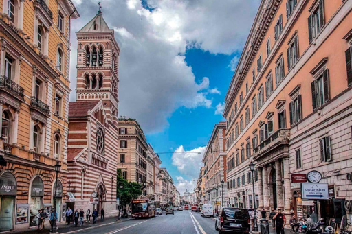 Fotografia panoramica di via Nazionale a Roma, ripresa dal centro della carreggiata in direzione del fondo della strada. Ai lati si affacciano edifici storici di diversi piani, con facciate color ocra, arancio e beige, finestre e negozi al piano terra. Sul lato sinistro si distingue un campanile con decorazioni in mattoni e pietra chiara. Sulla strada transitano autobus e automobili, mentre alcune persone camminano sui marciapiedi. Il cielo è parzialmente nuvoloso, con ampie zone azzurre.