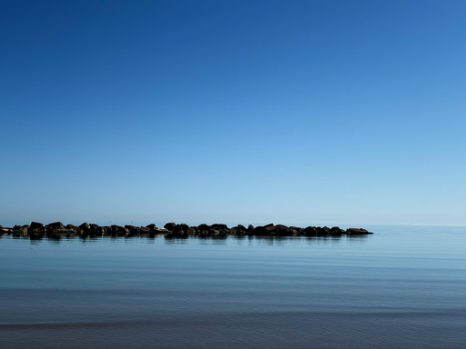 La fotografia mostra un mare calmo con acqua limpida e quasi priva di onde. In lontananza, si vede una fila di scogli che forma una barriera frangiflutti, separando il mare aperto dalla zona più vicina alla riva. Il cielo è completamente sereno e di un azzurro intenso, senza nuvole, creando un effetto di continuità tra mare e cielo. L’atmosfera è tranquilla e rilassante.