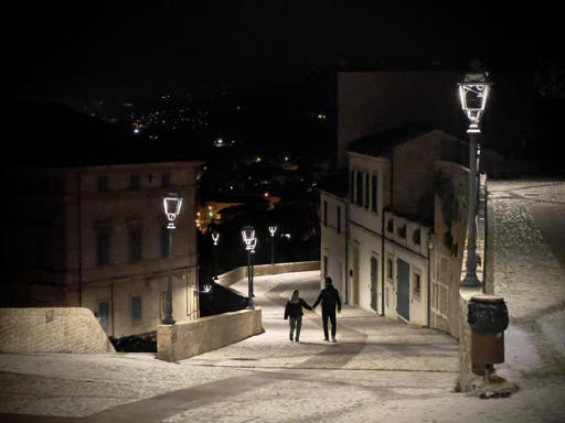 Due persone camminano a mano nella notte lungo una strada illuminate da lampioni (Grottammare -paese alto)