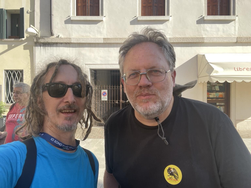 Un selfie di due persone all'aperto davanti a un edificio. La persona a sinistra (filippodb) ha capelli scuri lunghi e texturizzati, occhiali da sole scuri e indossa una maglietta azzurra con un cordino blu al collo. La persona a destra (Loris) ha capelli corti brizzolati, occhiali da vista e una maglietta nera con un piccolo logo circolare giallo e nero