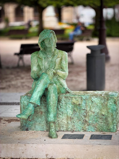 Statua di una donna seduta su un blocco, in bronzo patinato verde, in un parco all'aperto con panchine e persone sullo sfondo.
