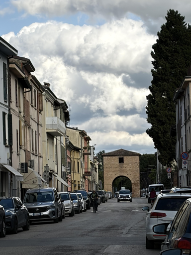 La foto mostra una strada urbana fiancheggiata da edifici di due o tre piani, in stile tradizionale, dai colori chiari e pastello, con persiane verdi e balconi. Le auto sono parcheggiate lungo entrambi i lati della via. Sullo sfondo si vede una porta medievale in mattoni con un grande arco centrale, oltre il quale si intravede un viale alberato. Il cielo è parzialmente nuvoloso, con nuvole bianche e grigie che lasciano filtrare un po’ di luce. A destra della porta si staglia un alto cipresso, che contribuisce al carattere pittoresco della scena.
