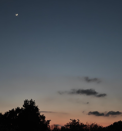Questa immagine cattura un momento di quiete al crepuscolo, dove i caldi colori del tramonto si fondono con l'oscurità della notte che avanza.
Ecco i dettagli principali:
Il Cielo: La parte superiore è dominata da un blu profondo e vellutato che sfuma gradualmente verso il basso in tonalità più chiare e polverose, fino a incontrare una striscia di arancione e rosa vibrante all'orizzonte.
La Luna: In alto a sinistra spicca una sottile falce di luna crescente, brillante e nitida contro il blu scuro. Poco più in basso, si nota anche un piccolo punto luminoso, probabilmente un pianeta o una stella particolarmente brillante.
Le Nuvole: Alcune nuvole scure e sfilacciate fluttuano a mezz'altezza, creando un contrasto materico con la fluidità del cielo.
La Silhouette: Lungo il bordo inferiore, le chiome degli alberi appaiono come sagome completamente nere, dando profondità alla scena e ancorandola alla terra.