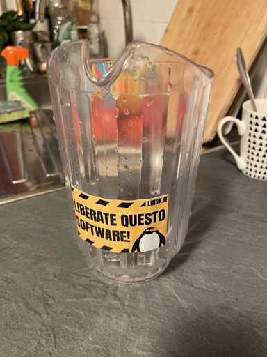 L'immagine mostra una caraffa trasparente in plastica posizionata su una superficie grigia, probabilmente una cucina. La caraffa ha un'etichetta gialla con bordi neri che recita "LIBERATE QUESTO SOFTWARE!" e "LINUX.IT", con un pinguino al centro. La caraffa ha condensa, suggerendo che è stata recentemente riempita con un liquido freddo. Sullo sfondo, si intravede un lavello con un rubinetto, un detergente verde e un asciugamano. A destra, c'è un bicchiere bianco con un motivo di triangoli neri. L'atmosfera è domestica e informale, con un tocco di humor e cultura open source.