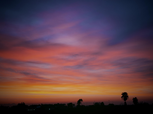 Colorful sunrise sky with shades of pink, purple, orange, and blue over a silhouette of trees and buildings.