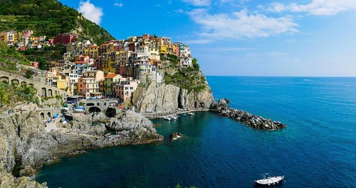 Fotografia che ritrae il borgo di manarola nelle Cinque Terre. Le case sono poste su una rupe in riva al mare, un molo di pietra protegge l'approdo, due barchette si dondolano in rada in una bella giornata di sole