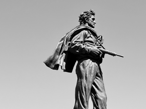 L’immagine mostra una statua in bronzo di un uomo in piedi. È il monumento al partigiano di Parma, con un mantello che gli ondeggia dietro come mosso dal vento. L’uomo indossa abiti semplici, con pantaloni e una camicia ampia, e tiene in mano un fucile puntato verso il basso. Il cielo sullo sfondo è completamente sgombro, creando un contrasto netto con la figura scura della statua.