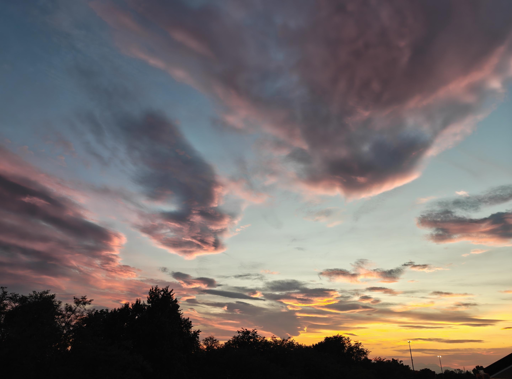 L'immagine mostra un tramonto spettacolare con tonalità vivide.Colori del CieloOrizzonte: Giallo intenso e arancione brillante.Cielo superiore: Sfumature azzurre e turchesi chiare.Nuvole: Toni rosa, viola e grigio scuro.Struttura delle NuvoleNuvole alte: Grandi formazioni soffici e piumate.Nuvole basse: Striature sottili e allungate vicino all'orizzonte.Contrasto: Forte stacco tra zone illuminate e ombre scure.Elementi in Primo PianoProfilo: Silhouette scura di alberi e vegetazione.Dettagli: Linea d'orizzonte completamente nera che esalta il cielo