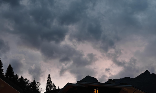 Il tramonto si nasconde dietro una fitta coltre di nuvole scure e cariche di pioggia, creando un'atmosfera drammatica e suggestiva sulle montagne di Alagna.Elementi visivi del cieloNuvole temporalesche: Masse grigie e bluastre dominano la parte superiore della scena.Sfumature rosa: Una luce tenue e rosata filtra dal basso, illuminando lo sfondo.Contrasto luminoso: Il chiarore crepuscolare crea un netto distacco con l'oscurità delle nubi.Il profilo del paesaggioSagome montuose: Le vette alpine si stagliano scure contro il cielo illuminato a contrasto.Alberi e tetti: I profili dei tetti delle case e delle cime dei pini incorniciano la parte inferiore.Luce calda: Una singola finestra illuminata emana una luce gialla che accentua il senso di rifugio