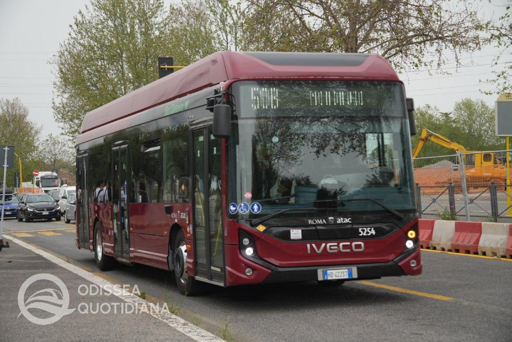 Un autobus elettrico Atac in servizio lungo una strada cittadina durante una giornata estiva. Il veicolo è fermo a una fermata con alcuni passeggeri in attesa, mentre sullo sfondo si notano edifici e alberature. L'immagine richiama il tema dell'introduzione della flotta elettrica e delle difficoltà operative legate alle temperature elevate, che incidono sull'autonomia delle batterie e sull'efficienza dell'impianto di climatizzazione.