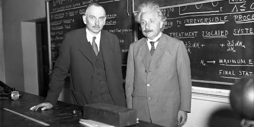 Fotografia in bianco e nero che ritrae Einstein davanti ad una lavagna piena di equazioni. Al suo fianco un altro fisico; entrambi vestiti formali con giacca e cravatta