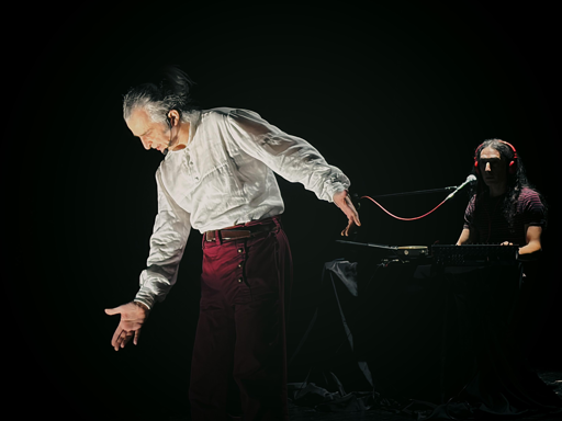 A performer in a white shirt gestures expressively, while a musician in the background plays a keyboard. The scene is set against a dark backdrop, emphasizing their dynamic interaction.