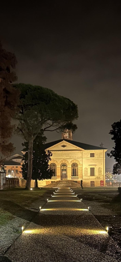 A well-lit pathway leads to a historic building at night, framed by trees and softly glowing lights. The architecture features classical elements, and the scene conveys a serene, atmospheric setting.