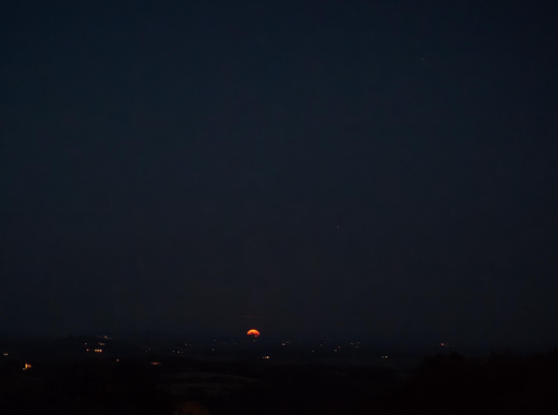 Questa fotografia notturna cattura il suggestivo momento in cui una luna calante, tinta di un profondo colore rosso-arancione, sorge o tramonta appena sopra la linea dell'orizzonte.
Ecco i dettagli principali dell'immagine:
Il cielo e i corpi celesti
La Luna: Posizionata in basso al centro, appare come un semicerchio luminoso e denso, parzialmente nascosto dal profilo terrestre. Il suo colore rosso intenso è tipicamente causato dalla rifrazione della luce attraverso l'atmosfera terrestre quando si trova molto bassa sull'orizzonte.
Le Stelle: Nel vasto cielo scuro e limpido che sovrasta la scena, si possono scorgere alcuni piccoli punti luminosi isolati che rappresentano le stelle o i pianeti più brillanti della notte.
Il paesaggio terrestre
L'orizzonte: Il profilo della terra è scuro e collinare, interrotto qua e là da piccole luci artificiali distanti che indicano la presenza di case, strade o piccoli centri abitati.
Primo piano: La parte inferiore dell'inquadratura è dominata da una fitta oscurità, dove si indovinano le sagome di alberi e vegetazione spontanea che incorniciano la vista panoramica