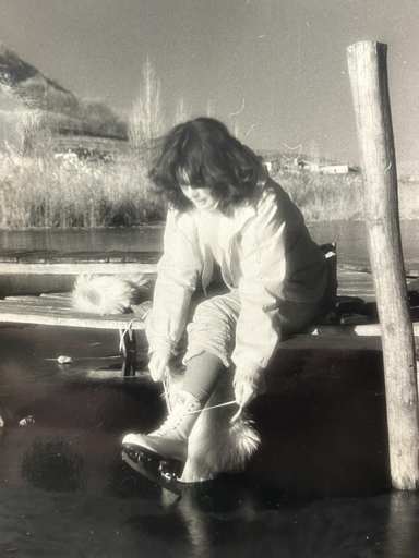 Foto in bianco e nero di una ragazza che si allaccia i pattini da ghiaccio seduta sul molo di un lago ghiacciato 