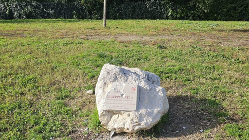 Un grande masso bianco con una targa commemorativa spaccata su di esso, situato in un'area verde con erba, sullo sfondo ci sono alberi e cespugli.