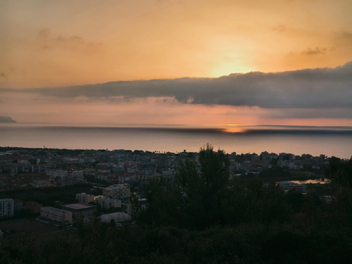 Un'alba arancione si riflette sul mare calmo oltre una città avvolta da una leggera foschia.